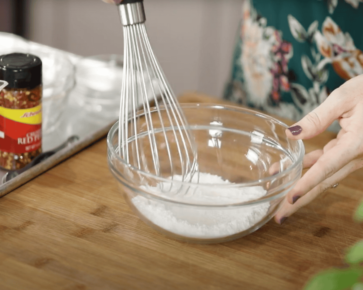 Whisking dry ingredients in a glass bowl on a wooden cutting board with red pepper flakes nearby
