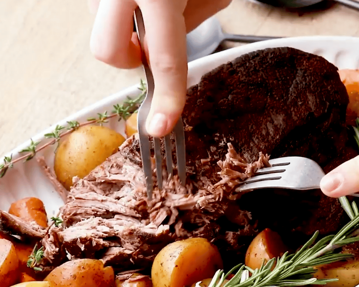 Forks shredding tender beef pot roast on a platter with cooked carrots, potatoes, and fresh herbs.