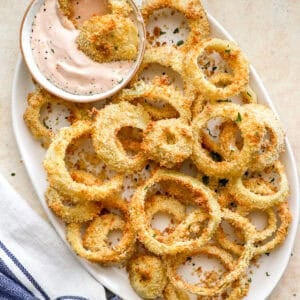 Golden, crispy air fryer onion rings served on a white plate with a bowl of creamy dipping sauce, sprinkled with herbs.