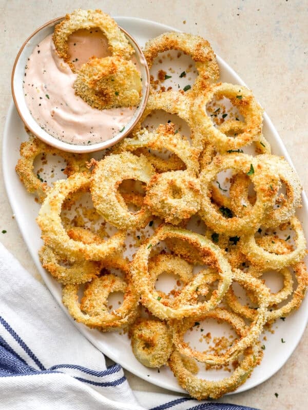 Golden, crispy air fryer onion rings served on a white plate with a bowl of creamy dipping sauce, sprinkled with herbs.