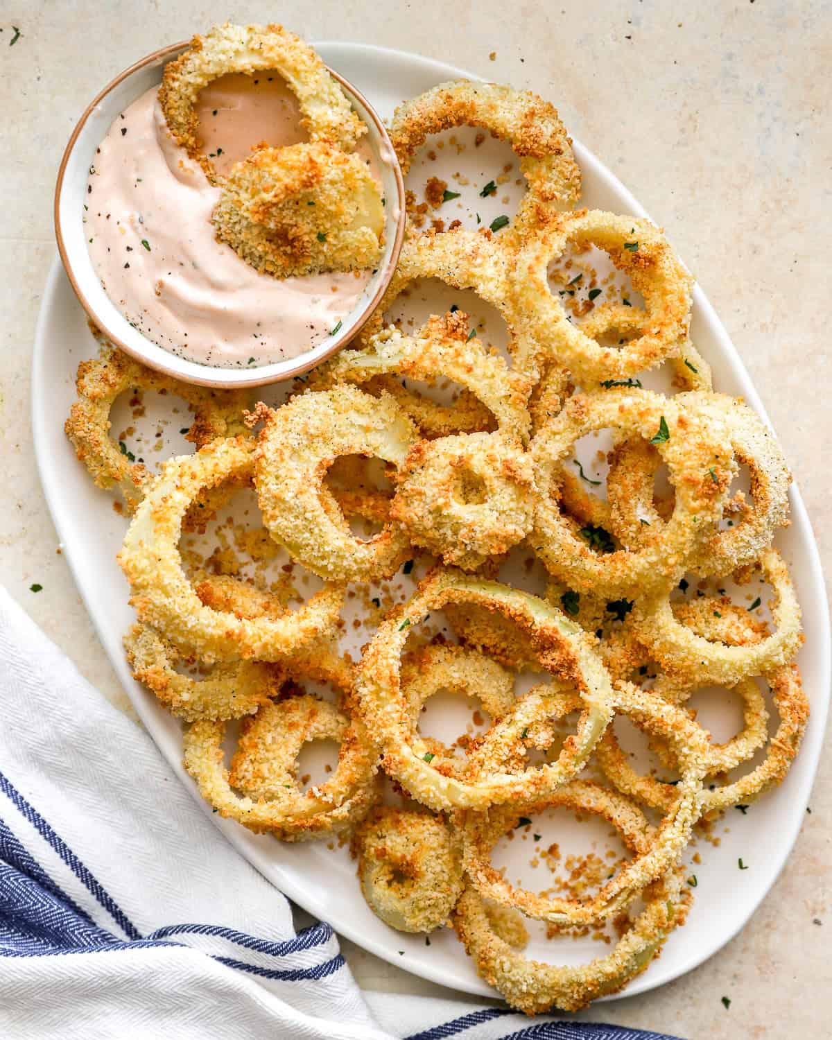 Golden, crispy air fryer onion rings served on a white plate with a bowl of creamy dipping sauce, sprinkled with herbs.