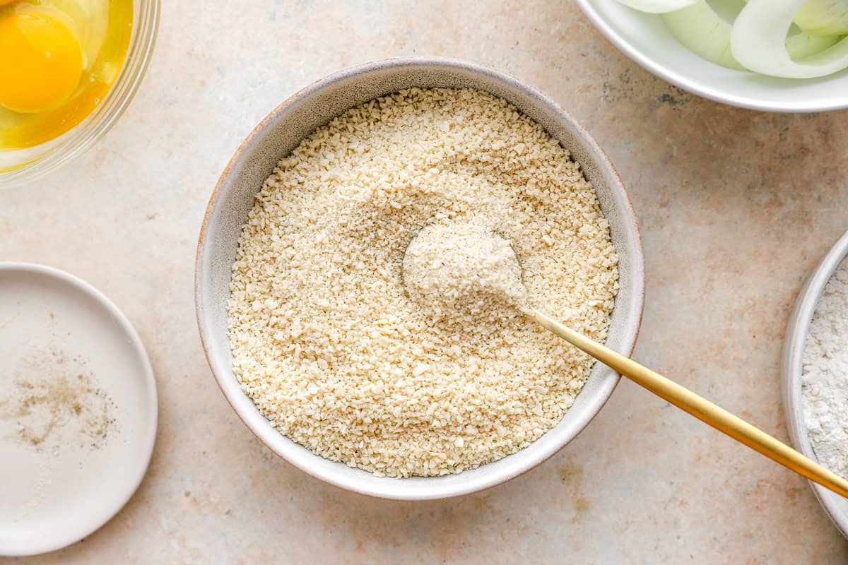 A bowl of seasoned panko breadcrumbs with a gold spoon, surrounded by small bowls of eggs, flour, and spices.