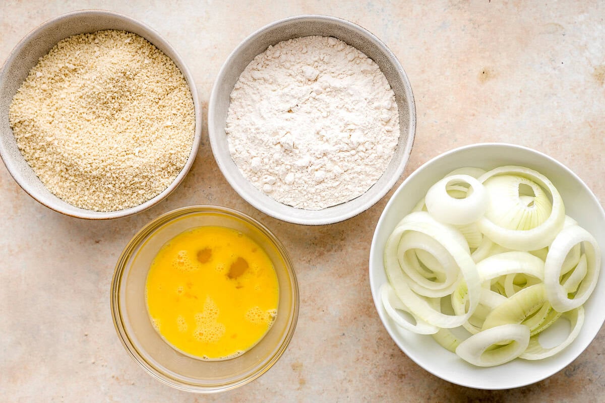 Bowls filled with panko breadcrumbs, flour, beaten eggs, and sliced raw onions arranged neatly on a countertop.