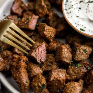 Close-up of juicy air fryer steak bites on a white plate with a side of creamy dipping sauce and a gold fork lifting a piece.
