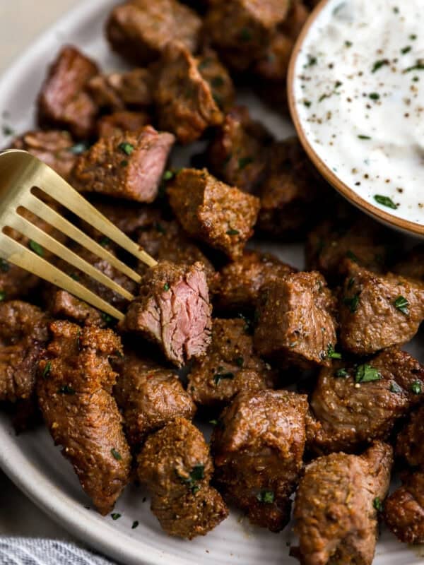 Close-up of juicy air fryer steak bites on a white plate with a side of creamy dipping sauce and a gold fork lifting a piece.