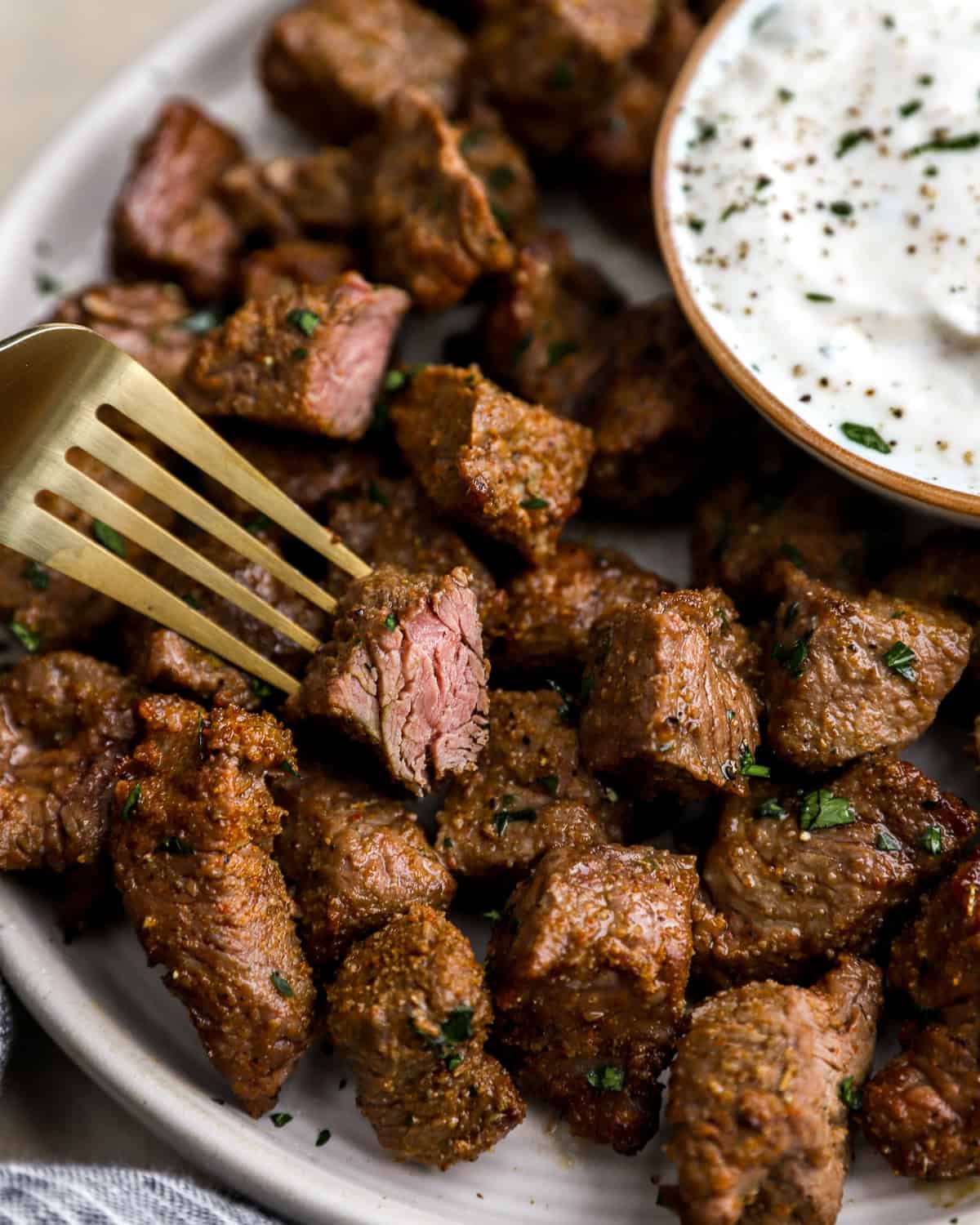 Close-up of juicy air fryer steak bites on a white plate with a side of creamy dipping sauce and a gold fork lifting a piece.
