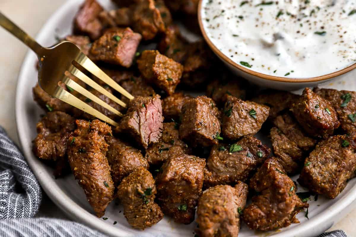 Fork picking up a tender, medium-cooked steak bite from a plate filled with seasoned steak pieces and sauce on the side.