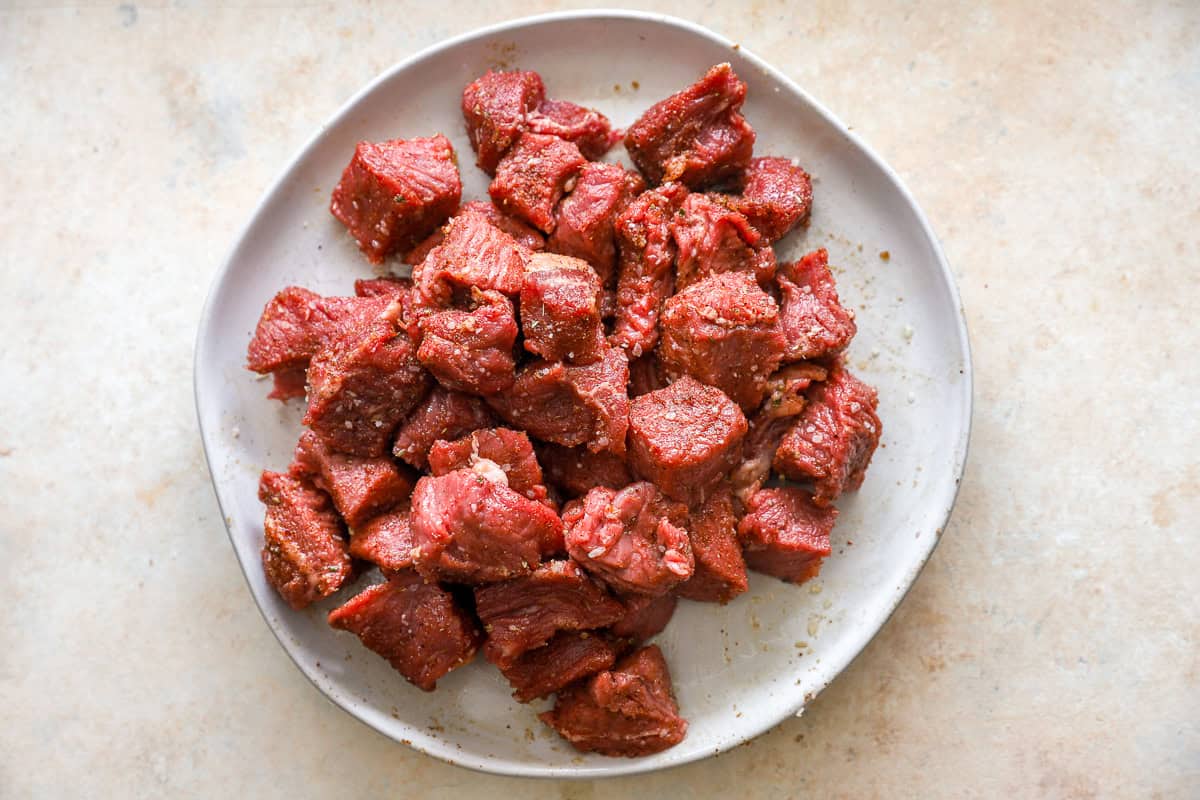 Raw steak cubes coated in the spice mix and olive oil on a white plate before air frying.