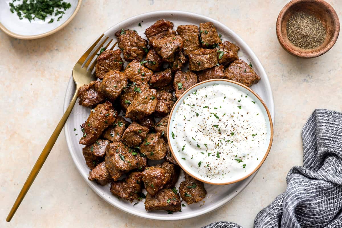 Plate of cooked air fryer steak bites garnished with parsley, served with a bowl of creamy dipping sauce.