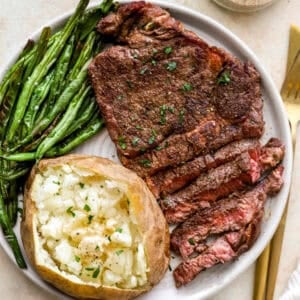 A juicy seasoned steak served with roasted green beans and a fluffy baked potato topped with butter and parsley on a white plate.