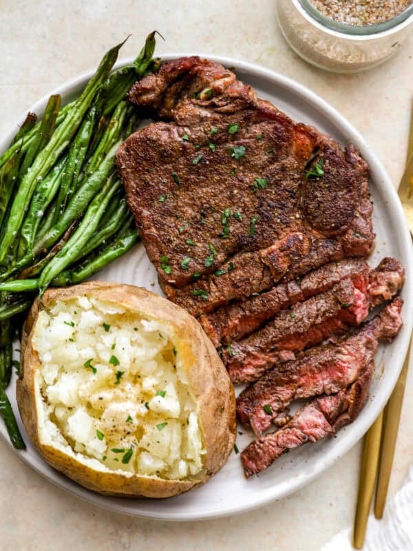 A juicy seasoned steak served with roasted green beans and a fluffy baked potato topped with butter and parsley on a white plate.