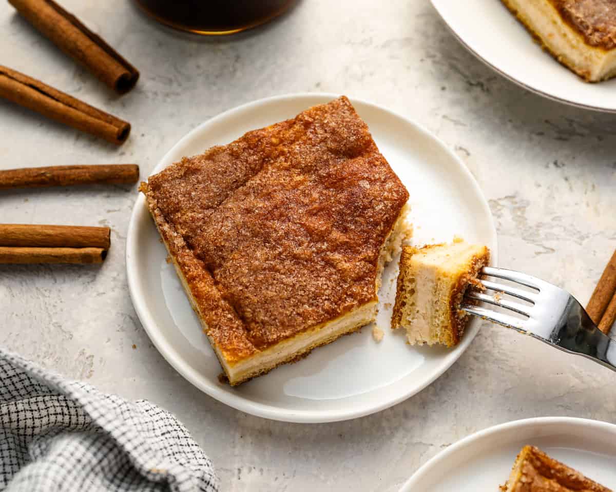 A slice of churro cheesecake on a white plate with cinnamon sticks around it and a fork taking a bite.