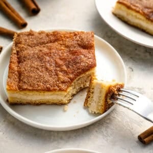 A slice of churro cheesecake on a white plate with a fork taking a bite, showing the creamy cheesecake layer and cinnamon sugar crust.
