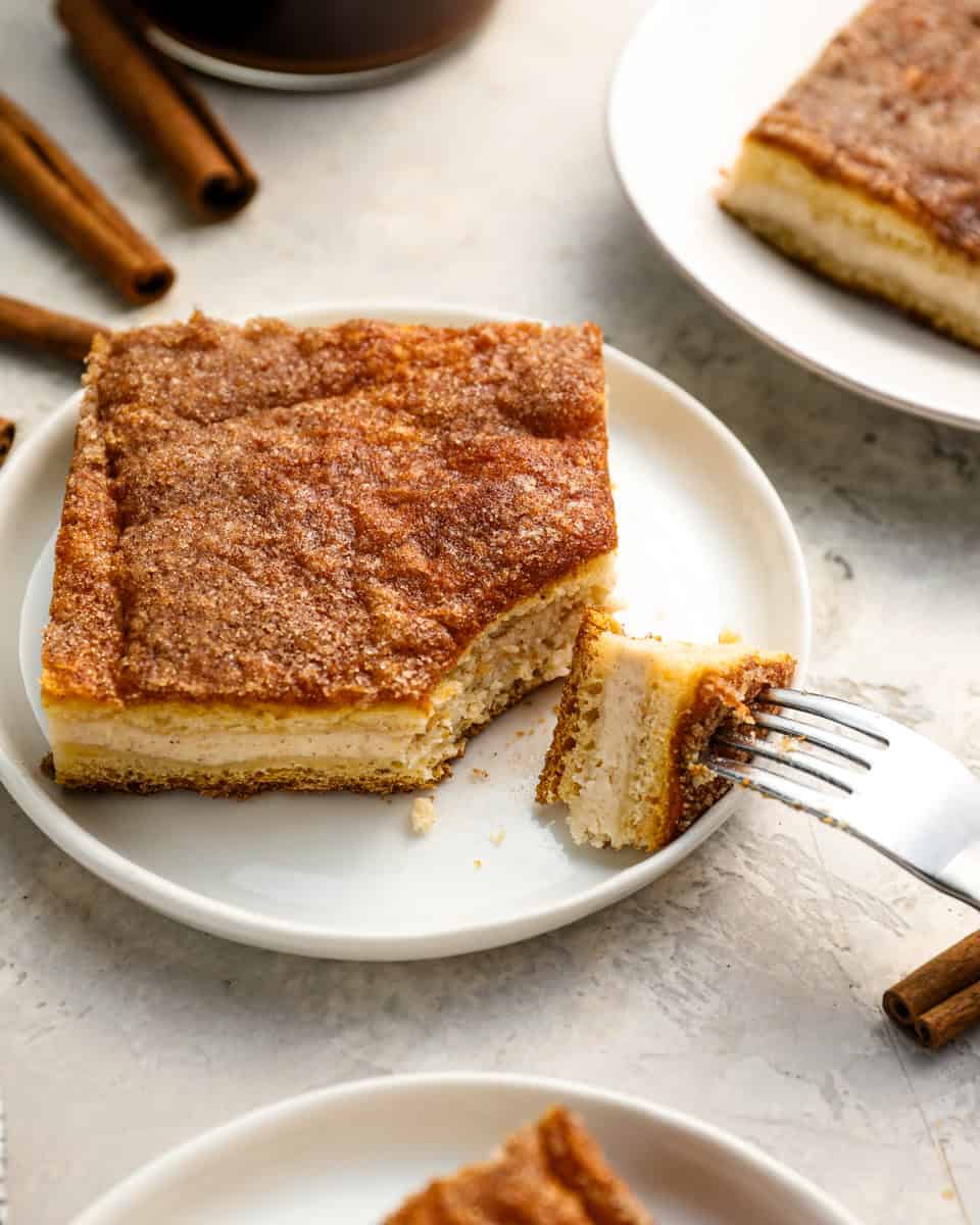 A slice of churro cheesecake on a white plate with a fork taking a bite, showing the creamy cheesecake layer and cinnamon sugar crust.
