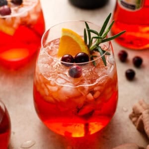 A cranberry Aperol spritz in a stemless glass filled with crushed ice, garnished with an orange slice, fresh cranberries, and a sprig of rosemary.