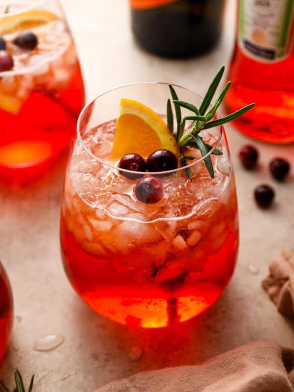 A cranberry Aperol spritz in a stemless glass filled with crushed ice, garnished with an orange slice, fresh cranberries, and a sprig of rosemary.