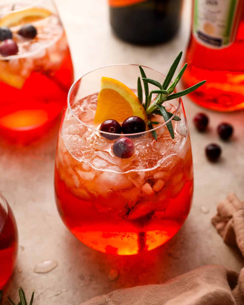 A cranberry Aperol spritz in a stemless glass filled with crushed ice, garnished with an orange slice, fresh cranberries, and a sprig of rosemary.