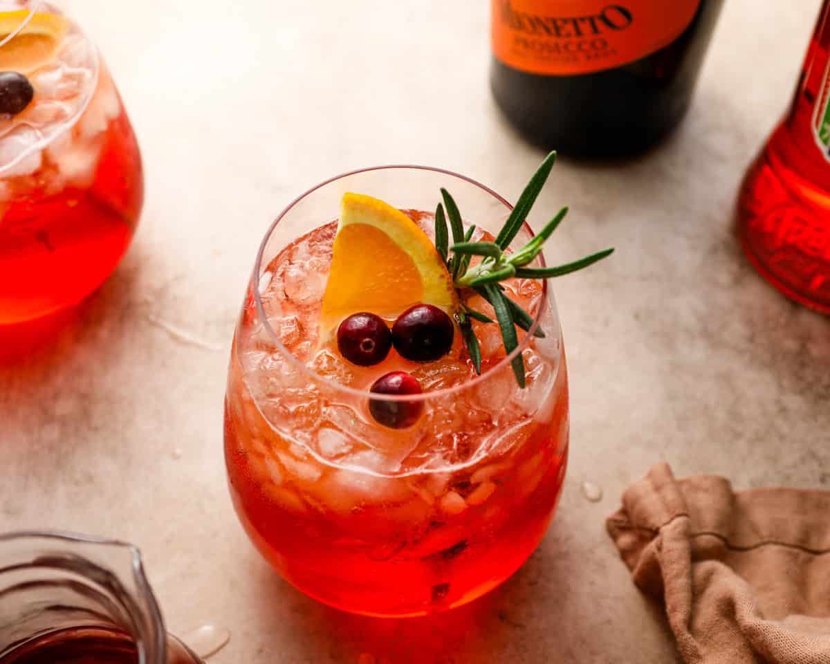 A cranberry Aperol spritz in a stemless glass with crushed ice, orange slice, cranberries, and rosemary, surrounded by bottles and holiday ingredients.