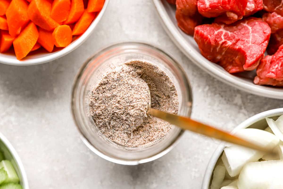 Overhead view of a jar filled with homemade beef stew seasoning mix surrounded by bowls of chopped carrots, raw stew meat, and diced onions.