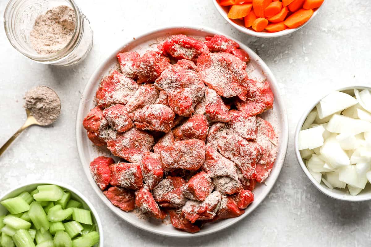 Raw beef stew meat coated in homemade beef stew seasoning mix on a plate, surrounded by bowls of chopped carrots, onions, and celery.