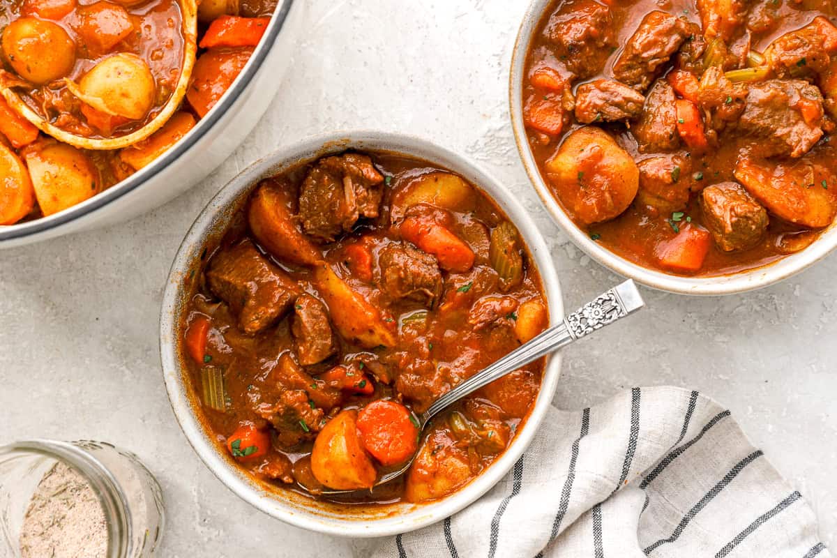 Three bowls of cooked beef stew with tender vegetables and chunks of beef in a thick, savory sauce, served with a striped kitchen towel.