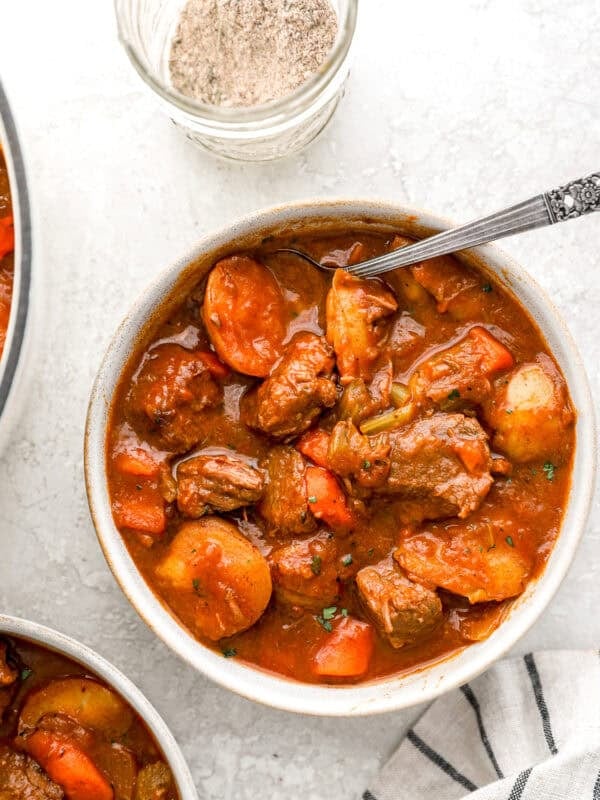 A bowl of hearty beef stew filled with tender beef chunks, carrots, potatoes, and celery in a rich broth, with a jar of homemade beef stew seasoning beside it.
