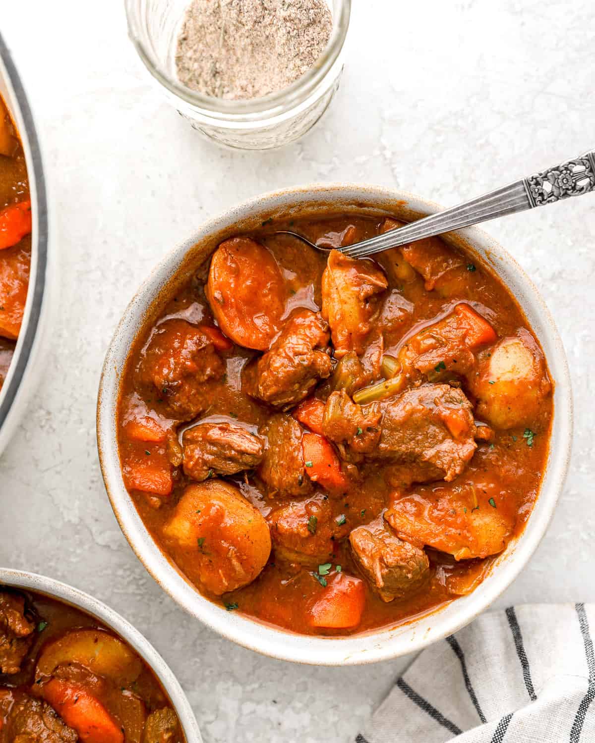 A bowl of hearty beef stew filled with tender beef chunks, carrots, potatoes, and celery in a rich broth, with a jar of homemade beef stew seasoning beside it.
