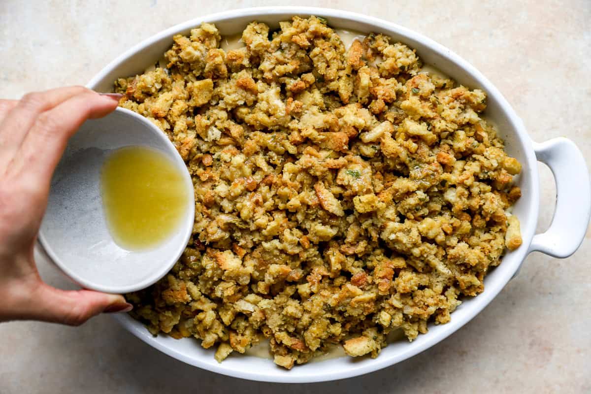Hand pouring melted butter over the top of the stuffing layer in the casserole before baking.