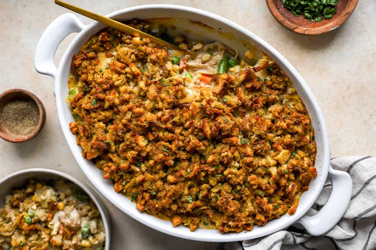 Golden baked chicken stuffing casserole in a white oval dish with a spoon and a serving scooped out, showing the creamy chicken and vegetable filling underneath.
