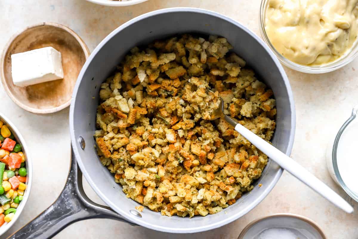 Prepared chicken stuffing mix in a saucepan with a spoon, surrounded by bowls of cream soup, milk, and frozen vegetables.