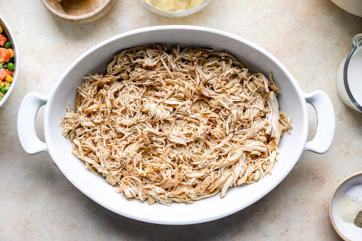 Shredded cooked chicken spread in an even layer in a white casserole dish, ready for the next layers of vegetables and sauce.