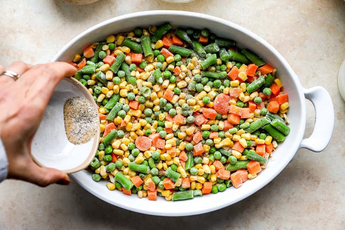 Hand sprinkling seasoning over a layer of frozen mixed vegetables on top of shredded chicken in a casserole dish.