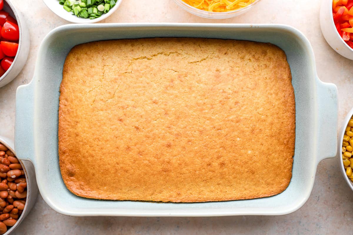 Freshly baked cornbread in a rectangular baking dish surrounded by small bowls of chopped vegetables, beans, cheese, and green onions.