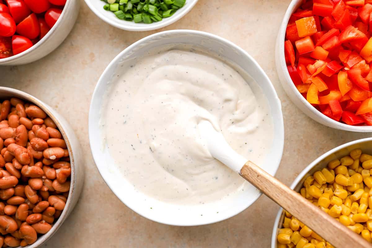 Bowl of homemade ranch dressing with a spatula, surrounded by bowls of chopped bell peppers, tomatoes, corn, beans, and green onions.