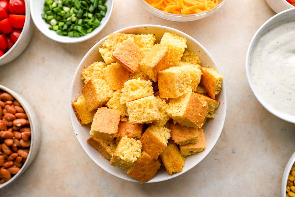 Bowl of sliced cornbread cubes ready for layering, with surrounding bowls of dressing, cheese, beans, and vegetables.