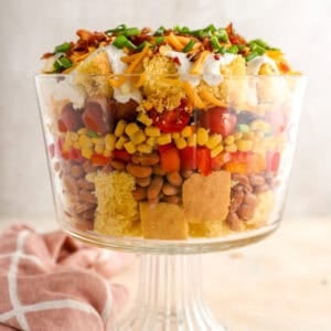Layered cornbread salad in a glass trifle dish with cornbread cubes, pinto beans, corn, tomatoes, peppers, bacon, cheddar cheese, and green onions.