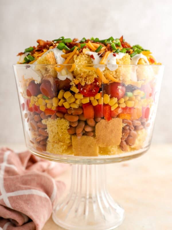 Layered cornbread salad in a glass trifle dish with cornbread cubes, pinto beans, corn, tomatoes, peppers, bacon, cheddar cheese, and green onions.