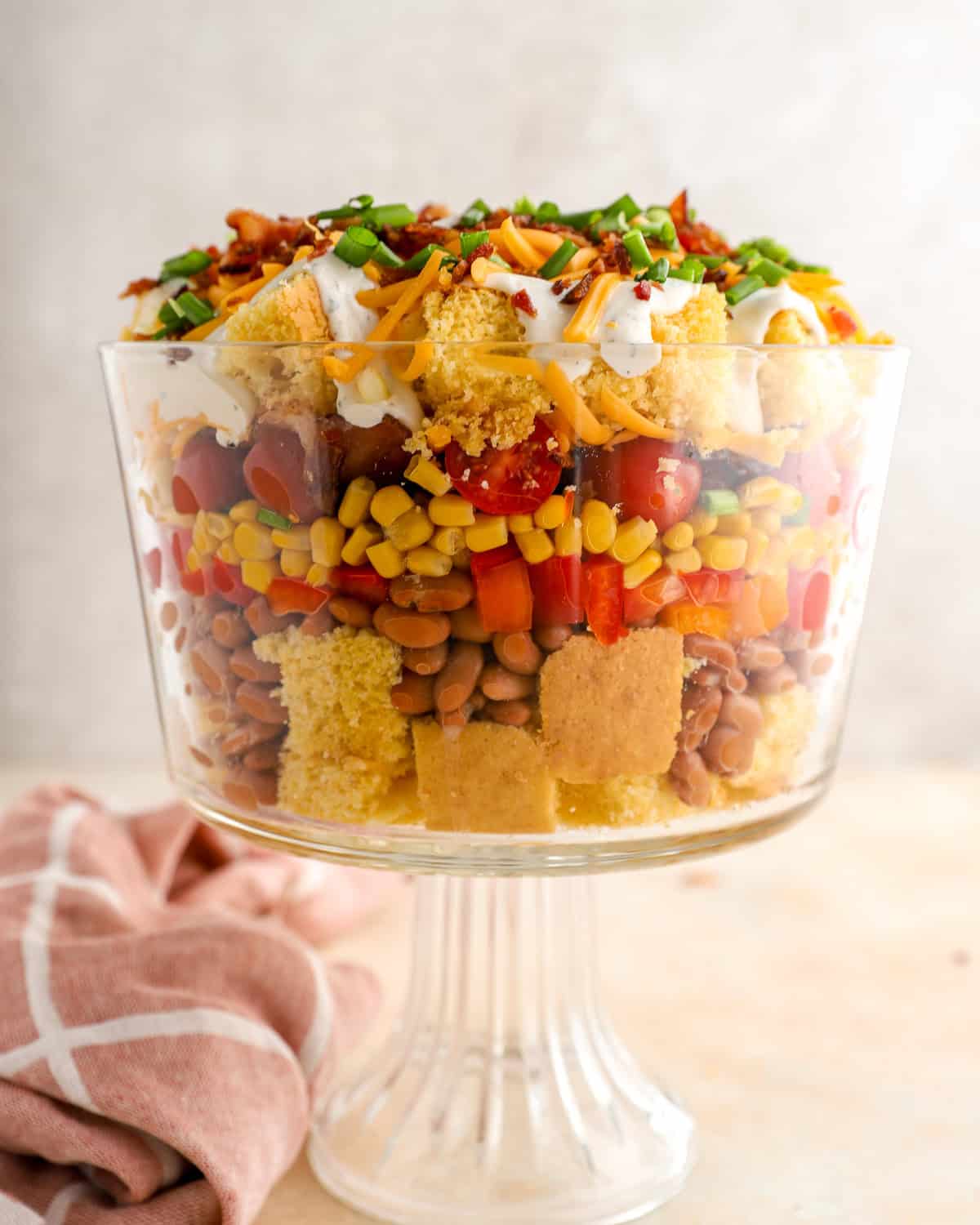 Layered cornbread salad in a glass trifle dish with cornbread cubes, pinto beans, corn, tomatoes, peppers, bacon, cheddar cheese, and green onions.