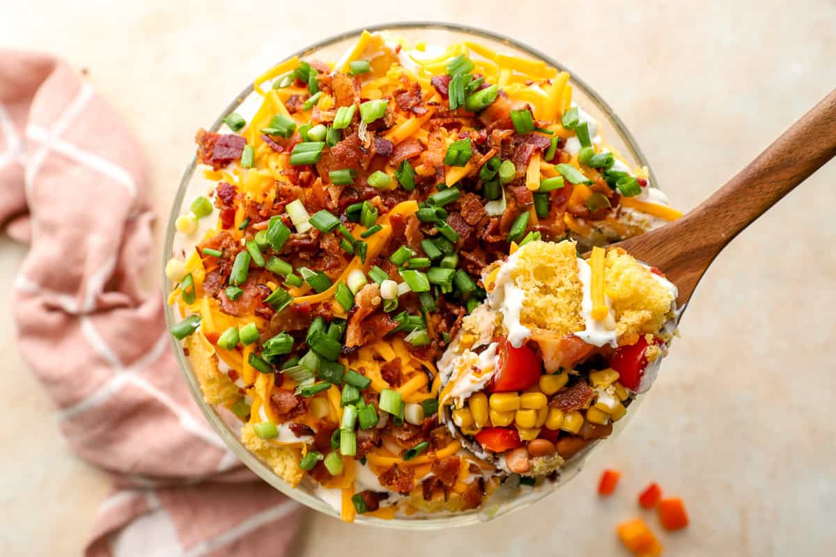Top view of cornbread salad with a wooden spoon scooping out layers of cornbread, corn, tomatoes, bacon, and creamy dressing.