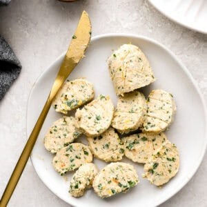 Slices of chilled cowboy compound butter on a white plate with a gold butter knife.