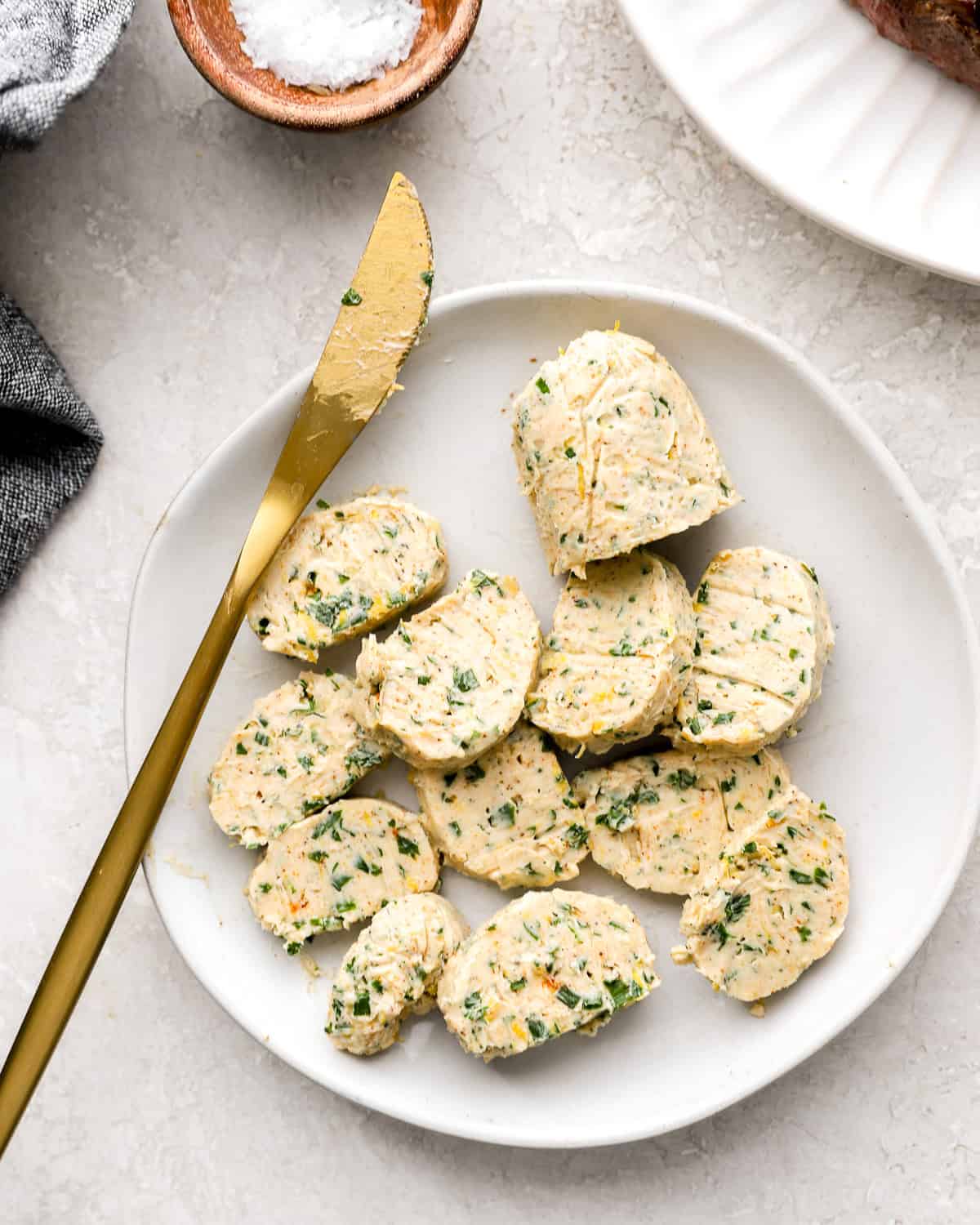 Slices of chilled cowboy compound butter on a white plate with a gold butter knife.