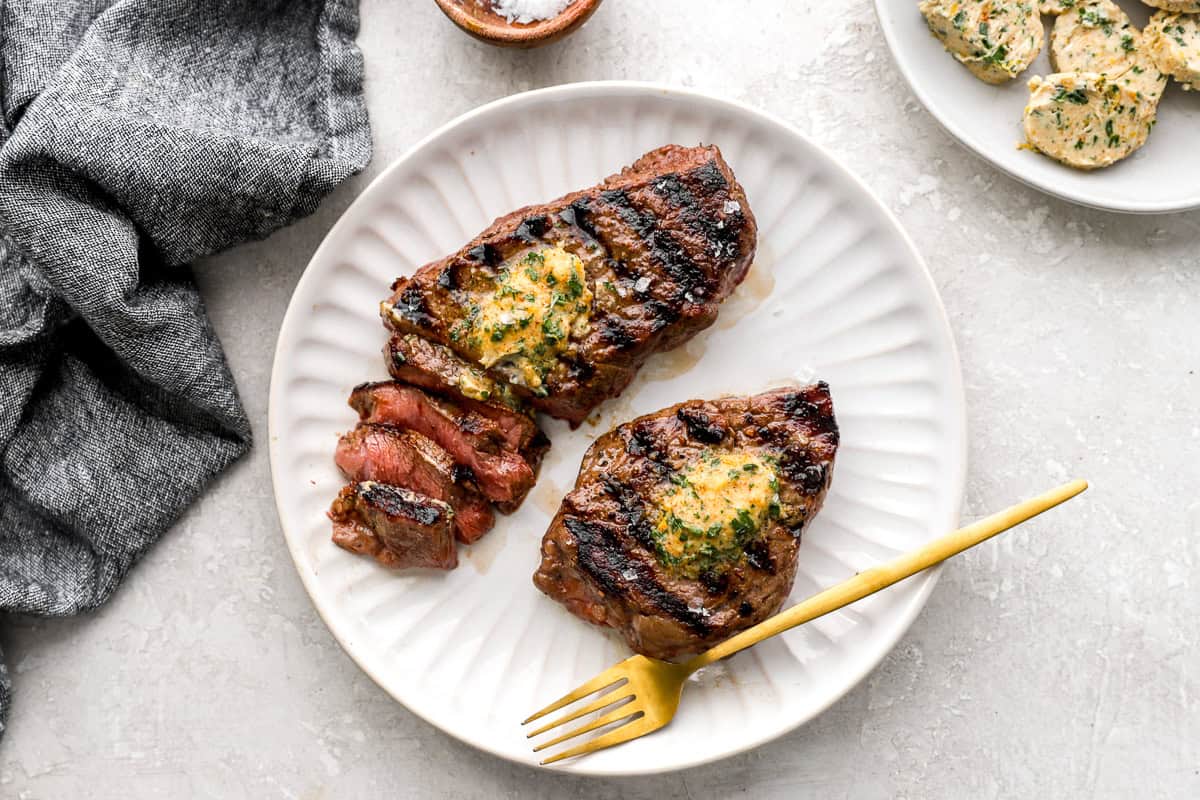 Grilled steaks topped with melted cowboy butter on a white plate with a gold fork.