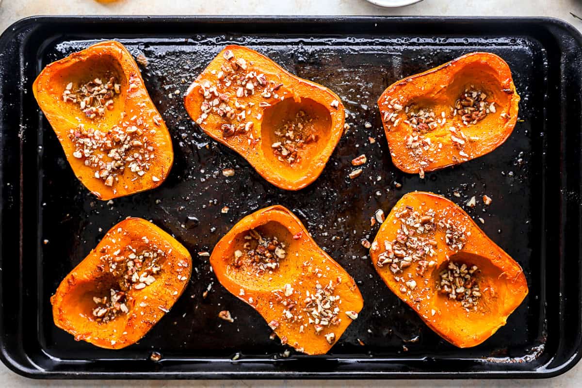 Roasted honeynut squash halves on a baking sheet topped with chopped pecans before finishing in the oven.