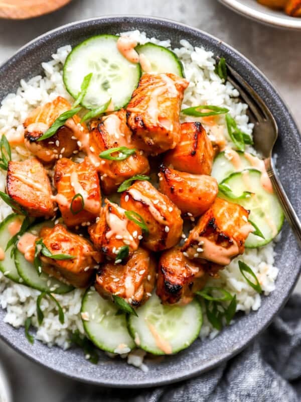 A bowl of air fryer salmon bites served over white rice with sliced cucumbers, sesame seeds, green onions, and sriracha mayo drizzled on top.