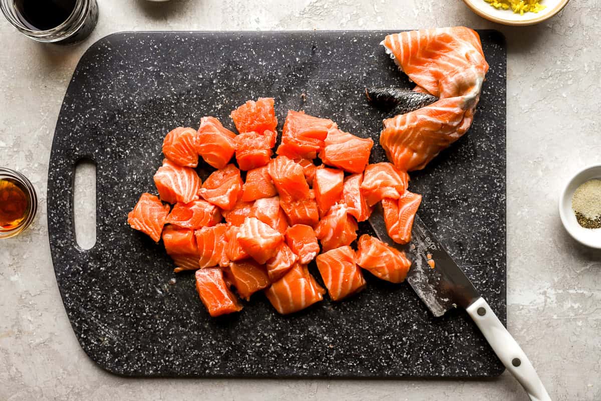 Raw salmon fillet cut into chunks on a black cutting board, with the salmon skin and a knife resting beside it.
