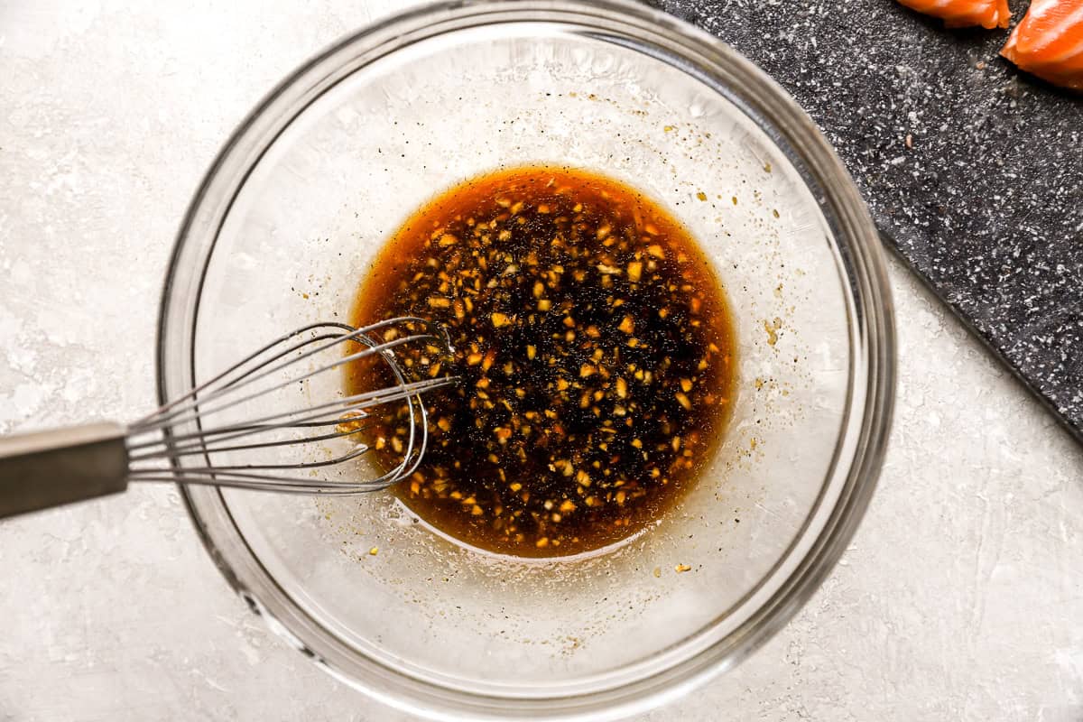 A glass mixing bowl filled with soy sauce, honey, garlic, ginger, and sesame oil marinade with a whisk inside.