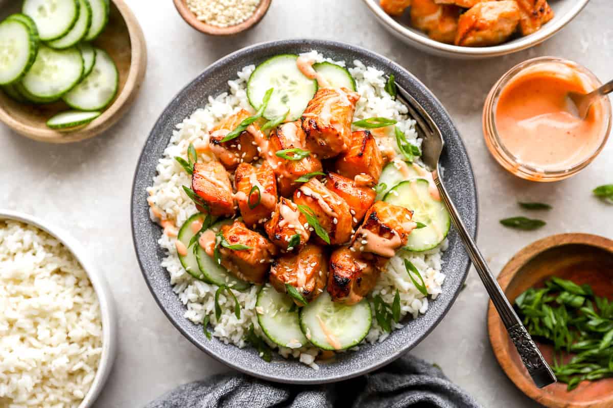 A plated salmon bowl with rice, cucumber slices, sriracha mayo, green onions, and sesame seeds, surrounded by small bowls of toppings.