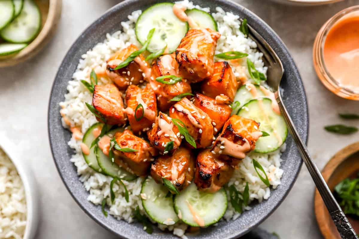 Close-up overhead view of caramelized salmon bites over rice with cucumbers, sesame seeds, green onions, and spicy mayo drizzle.