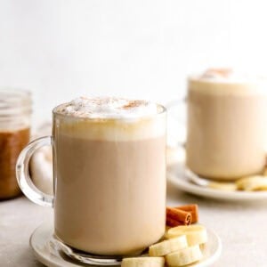Banana bread latte topped with frothy milk and cinnamon, served in a glass mug with sliced banana and cinnamon sticks