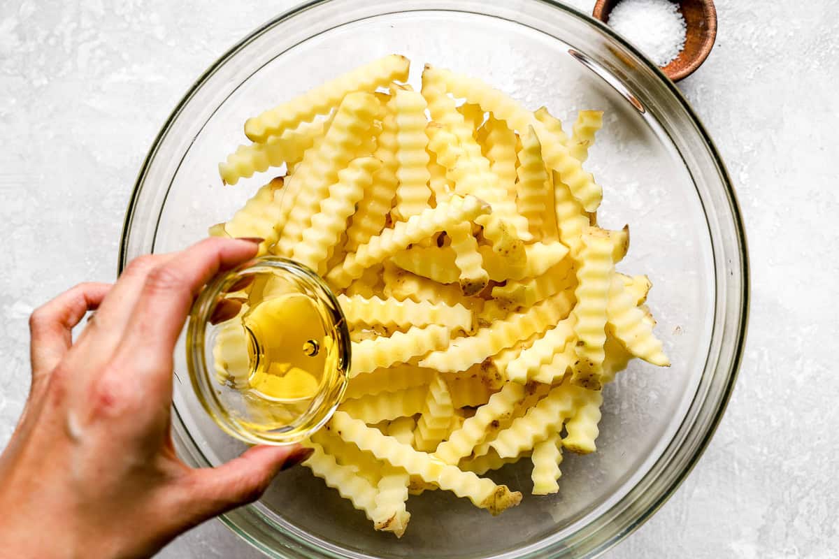 Olive oil being poured over crinkle cut potatoes in a glass bowl before seasoning
